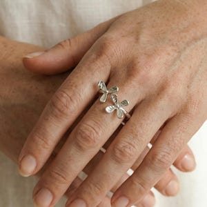 May include: A silver ring featuring a floral design with two flower-shaped elements. The ring is worn on a finger, showcasing the intricate details of the petals. The jewelry is made of a shiny, metallic material.
