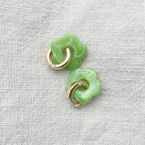 Könnte beinhalten: Ein Paar grüne und goldene Ohrringe in Blumenform. Die Ohrringe sind aus hellgrünem Kunststoff mit einem Wirbelmuster und haben einen goldenen Reifen.