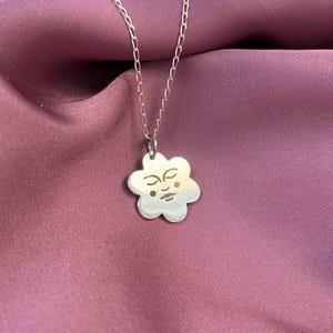 Könnte beinhalten: Eine silberne Kettenkette mit einem silbernen Blumenanhänger. Die Blume hat ein Gesicht mit geschlossenen Augen und einem kleinen Mund.