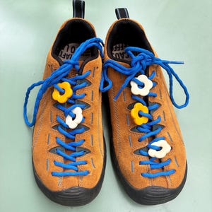 May include: A pair of tan suede trainers with bright blue laces and black rubber soles. Each shoe features three small, colourful flower-shaped charms threaded onto the laces, with yellow and white petals visible on the front of the footwear.