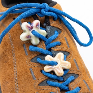 May include: A brown suede shoe with bright blue laces threaded through two ceramic flower-shaped charms. The top charm is cream with purple accents, while the bottom charm is a solid light beige. The shoe features dark grey stitching.