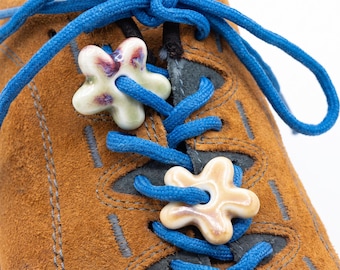May include: A brown suede shoe with bright blue laces threaded through two ceramic flower-shaped charms. The top charm is cream with purple accents, while the bottom charm is a solid light beige. The shoe features dark grey stitching.