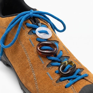 Puede incluir: Una zapatilla de ante marrón con cordones azul brillante enhebrados a través de tres dijes decorativos ovalados. Los dijes están hechos de material translúcido en colores transparente, ámbar y verde oscuro, cada uno con un orificio.
