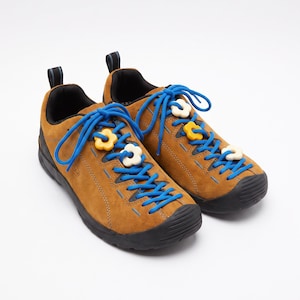 Puede incluir: Un par de zapatillas de ante marrón con cordones azul brillante y suelas de goma negra. Los cordones están decorados con pequeños abalorios en forma de flor de color amarillo y blanco roto. Cada zapato tiene una lengüeta negra en el talón.