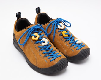 May include: A pair of brown suede sneakers with bright blue laces and black rubber soles. The laces are decorated with small, colorful flower-shaped charms in yellow and off-white. Each shoe features a black pull tab at the heel and a rounded toe.