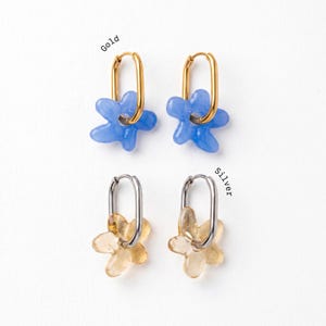 May include: Two pairs of hoop earrings with flower-shaped charms. The top pair features gold-tone oval hoops with blue flower charms labelled Gold. The bottom pair features silver-tone oval hoops with translucent yellow flower charms labelled Silver.