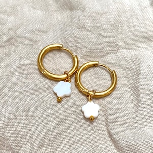 Peut inclure: Une paire de créoles dorées avec un charme de fleur blanche suspendu à chaque créole.