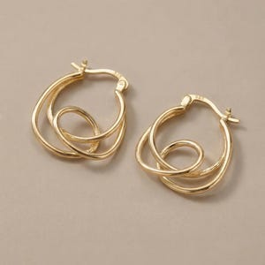 Puede incluir: Un par de pendientes de aro dorados. Cada pendiente presenta un diseño único con bucles entrelazados, creando un aspecto elegante y moderno. Los pendientes están sobre un fondo neutro.