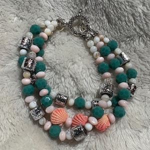 May include: A three-strand bracelet featuring teal, white, and pink beads, interspersed with silver-toned decorative elements and small coral-colored seashell charms. The bracelet is secured with a silver toggle clasp.