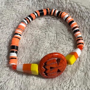 May include: A Halloween-themed bracelet featuring orange, black, and white disc beads. The bracelet includes a carved orange pumpkin bead and candy corn accents, perfect for the spooky season.
