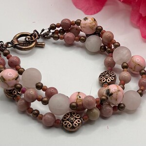 May include: A multi-strand bracelet featuring pink and white beads with copper-colored accents. The bracelet has a toggle clasp and decorative metal beads. The beads vary in size and texture, creating a layered effect.