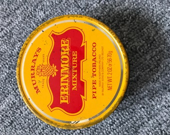 Op de afbeelding: Een gele en rode blik met de tekst "MURRAYS TRADE MARK" en "ERINMORE MIXTURE PIPE TOBACCO". De blik wordt gemaakt in Noord-Ierland door Murray Sons & Company Ltd Belfast. Het nettogewicht is 2 oz of 56-70g.