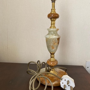 May include: A vintage table lamp with a marble base and a brass stem. The lamp features a decorative, ornate design with a light green and brown marble body. The electrical cord is coiled on the table.