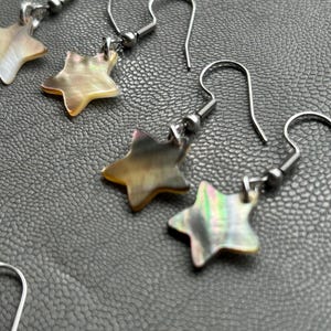 May include: Earrings with star-shaped pendants. The pendants have a pearlescent, iridescent finish with shades of gold, brown, and green. The earring hooks are silver-colored.