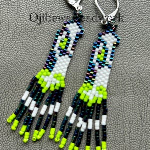 May include: Handmade beaded earrings with a unique design. The earrings feature a white base with neon green, black, and iridescent accents. The fringe is neon green and black. The earrings have silver-tone lever-back closures. The text "O jibewa beadwork" is visible.