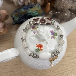 Może przedstawiać: Bransoletka z koralików z centralną cylindryczną koralikiem z brązowym i białym wzorem. Bransoletka zawiera okrągłe, jasnoróżowe koraliki i dekoracyjne srebrne metalowe akcenty. Z bransoletki zwisa mały, złoty urok.