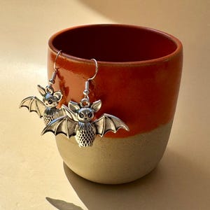 May include: A pair of silver-toned bat-shaped earrings with detailed wing textures and small ears, hanging from standard metal hooks. The earrings are displayed resting against the rim of a two-toned ceramic cup with a glossy orange top and matte beige base.