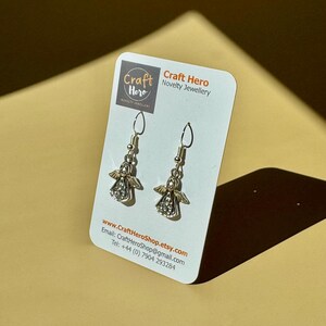 May include: A pair of silver-toned angel earrings with rhinestone accents, displayed on a white card with the text "Craft Hero Novelty Jewellery." The earrings have a detailed design and are suspended from silver hooks. The card also includes the website and contact information.