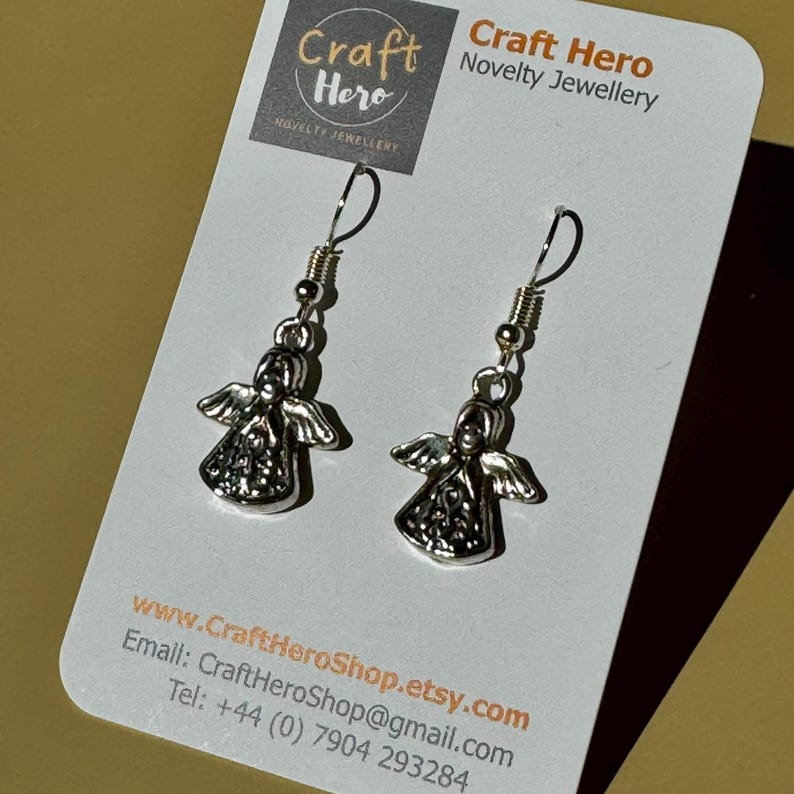 May include: A pair of silver-colored angel earrings with detailed wings and a small head. The earrings hang from silver-colored hooks. The earrings are displayed on a white card with the text "Craft Hero Novelty Jewellery".