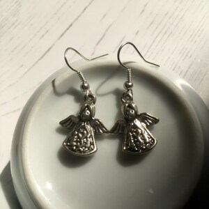 May include: A pair of silver-toned angel earrings. Each earring features a small angel pendant with outstretched wings and intricate detailing. The earrings have a simple hook closure.