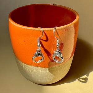 May include: A pair of silver-toned metal dangle earrings featuring miniature handcuff charms. Each earring is suspended from a standard curved ear wire hook, showcasing a polished metallic finish and detailed circular links that resemble functional handcuffs.