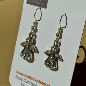 May include: A pair of silver-toned angel earrings. Each earring features a small angel charm with detailed wings and a decorative pattern on the body. The earrings hang from a simple hook and coiled wire design.