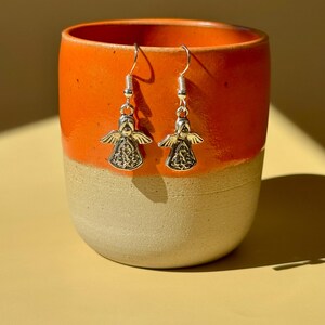 May include: A pair of silver angel earrings with detailed wings and a decorative design on the body. The earrings are hanging in front of an orange and beige ceramic mug.