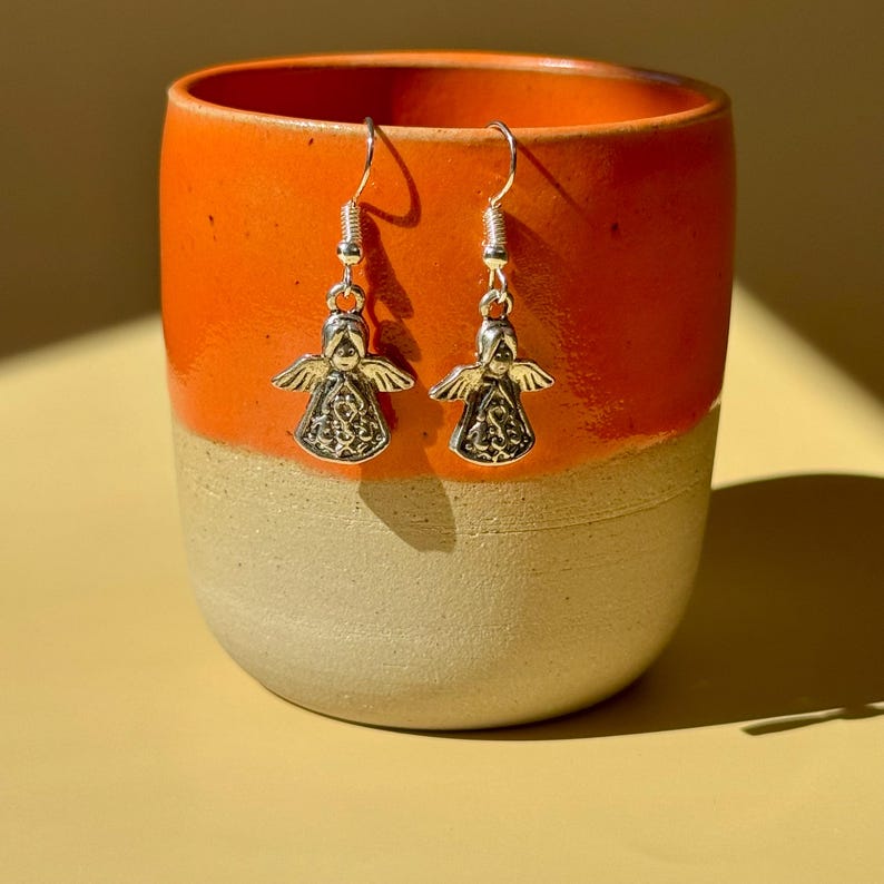 May include: A pair of silver angel earrings with detailed wings and a decorative design on the body. The earrings are hanging in front of an orange and beige ceramic mug.