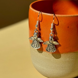 May include: A pair of silver-colored angel earrings with detailed wings and robes. The earrings are hanging in front of an orange and beige ceramic mug, creating a warm, inviting composition. The earrings are a delicate accessory.