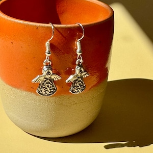 May include: A pair of silver angel earrings with detailed wings and dresses. The earrings hang from silver hooks and are displayed in front of an orange and beige ceramic mug. The earrings are a religious or spiritual themed jewelry item.