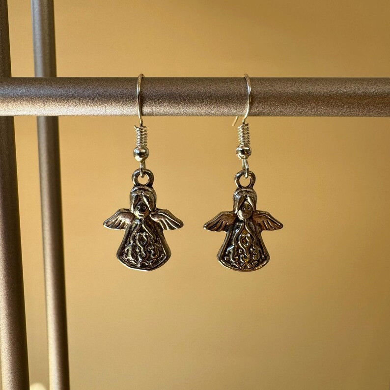 May include: A pair of silver-toned angel earrings. Each earring features a detailed angel pendant with outstretched wings and intricate detailing. The earrings hang from silver-colored fish hook ear wires.