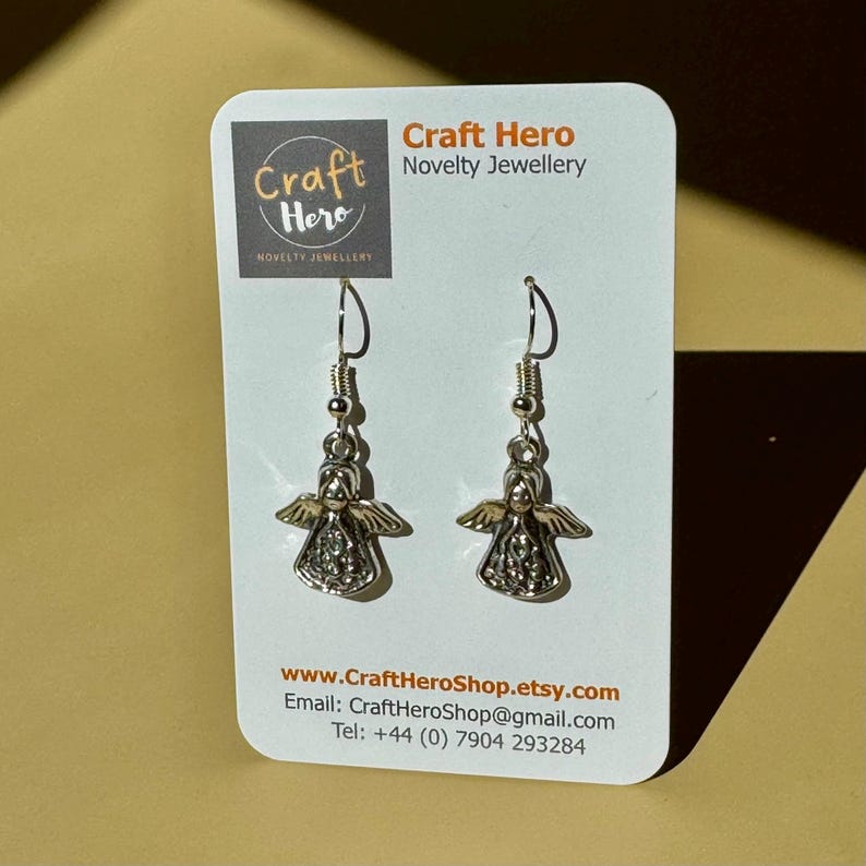 May include: Silver-toned angel earrings with detailed wings and a decorative design. The earrings are displayed on a white card with the "Craft Hero Novelty Jewellery" logo and contact information.