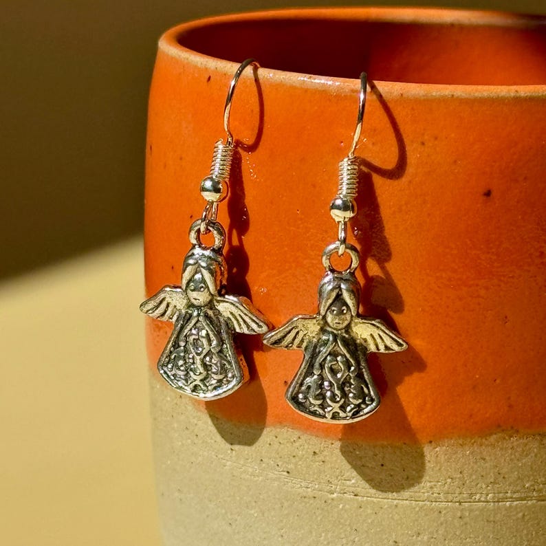 May include: A pair of silver-toned angel earrings. Each earring features a detailed angel pendant with outstretched wings and a decorative dress. The earrings hang from a simple hook.
