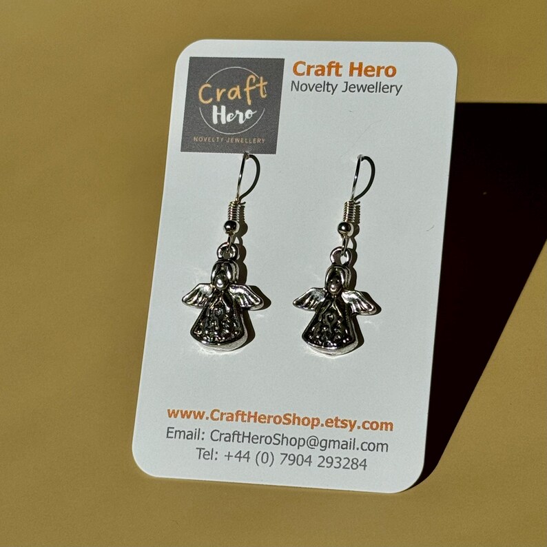 May include: A pair of silver-colored angel earrings with detailed wings and bodies, displayed on a white card with the Craft Hero Novelty Jewellery logo and contact information.