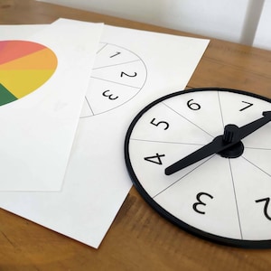 May include: A white game spinner with black numbers and a black arrow, next to two white papers. One paper has a color wheel, the other has a numbered circle. The items are on a wooden surface.