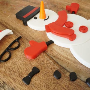 May include: A disassembled snowman kit with white body pieces, a black hat, and red accessories. Includes a carrot nose, black glasses, a bow tie, and coal-like pieces for the mouth. The pieces are arranged on a wooden surface.