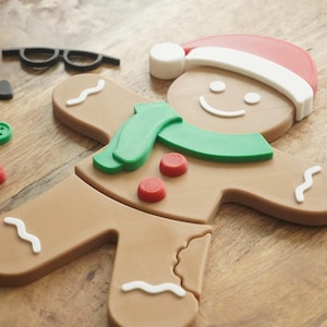 May include: A gingerbread man Christmas decoration with a red and white Santa hat, green scarf, and red buttons. Also includes a black bow tie, glasses, and a green button. The gingerbread man is on a wooden surface.