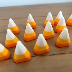 May include: A collection of thirteen small, triangular candy corn-shaped objects. Each piece features a distinct three-color horizontal striped pattern consisting of a white top section, a yellow middle section, and an orange base section.