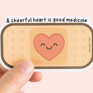 May include: A sticker featuring a bandage design with a smiling heart in the center. The text "A cheerful heart is good medicine" is at the top, with "PROVERBS 17:22" on the right side. The sticker is held by a hand.