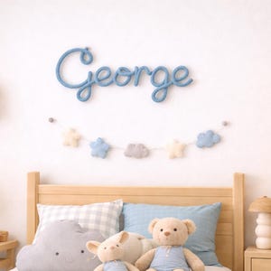May include: A blue cursive "George" wall decoration above a bed with a wooden headboard. A garland of felt stars and clouds hangs below the name. The bed has pillows and soft toys. A wooden shelf and bedside table are visible.