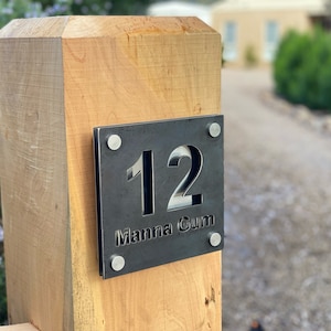 May include: A black metal house number sign with the number 12 and the text "Manna Gum" attached to a wooden post.
