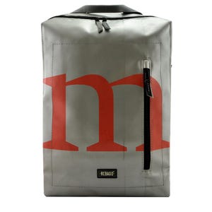 May include: A rectangular silver backpack featuring a large red lowercase letter m printed on the front. The bag includes a vertical black zippered pocket on the right side, a black top handle, and a small black rectangular label with the text REBAGO.