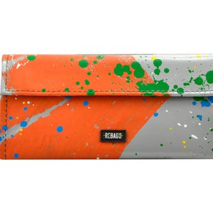 May include: A grey and orange wallet with a splatter paint design. The wallet has a black label that says "REBAGCO".