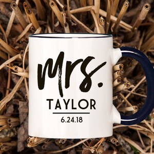 May include: White ceramic mug with a black rim and a navy blue handle. The mug has black text that says "Mrs. TAYLOR 6.24.18".