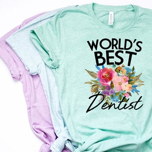 Puede incluir: Tres camisetas apiladas en tonos morado, azul claro y verde menta. La camiseta verde menta superior presenta el texto "WORLD'S BEST Dentist" sobre un ramo floral con flores rosas, azules y moradas.