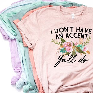 以下が含まれることがあります： 「I don't have an accent y'all do」というテキストと花柄のデザインが施されたピンクのTシャツです。