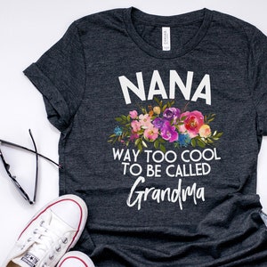 Puede incluir: Una camiseta gris oscuro con un dise&ntilde;o floral blanco y el texto "NANA WAY TOO COOL TO BE CALLED GRANDMA".