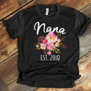 Puede incluir: Una camiseta gris oscuro con un dise&ntilde;o floral blanco y el texto "Nana Est. 2018".