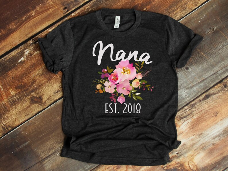 Puede incluir: Una camiseta gris oscuro con un dise&ntilde;o floral blanco y el texto "Nana Est. 2018".