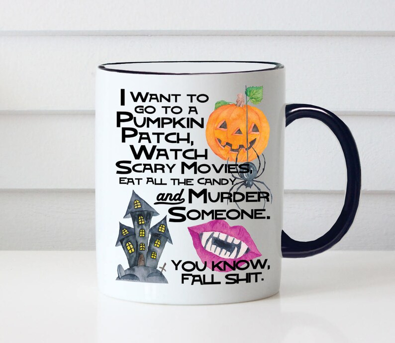 May include: White ceramic coffee mug with a black rim and handle. The mug features a Halloween-themed design with a pumpkin, a haunted house, and a pair of lips with fangs. The text on the mug reads "I want to go to a pumpkin patch, watch scary movies, eat all the candy and murder someone. You know, fall shit."